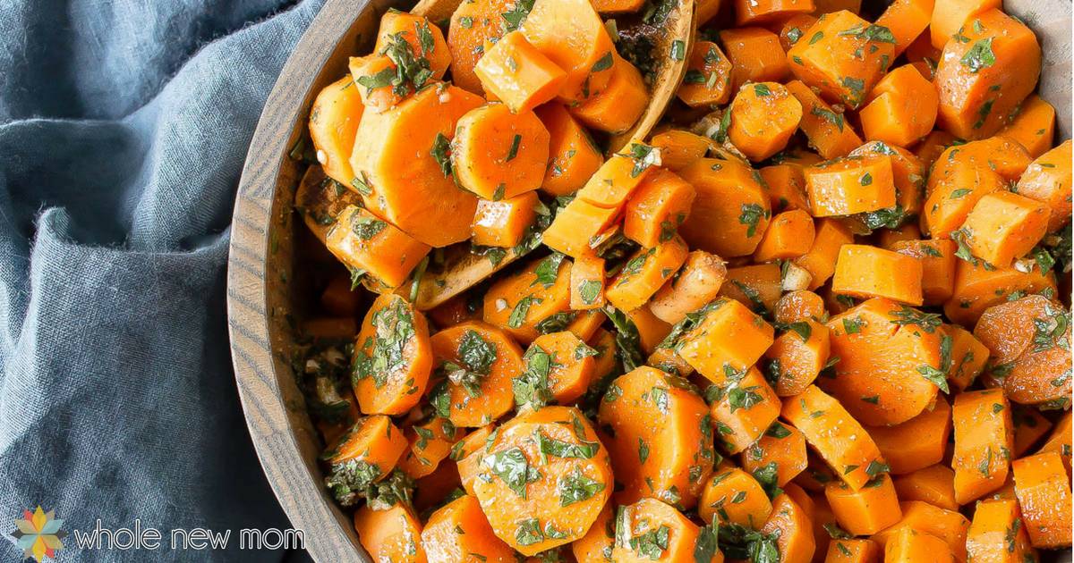 A vibrant dish of diced and sliced cooked carrots garnished with chopped herbs sits in a wooden bowl. The background features a blue cloth draped casually, adding contrast. The image branding includes the text “whole new mom” with a small flower logo in the bottom left corner.