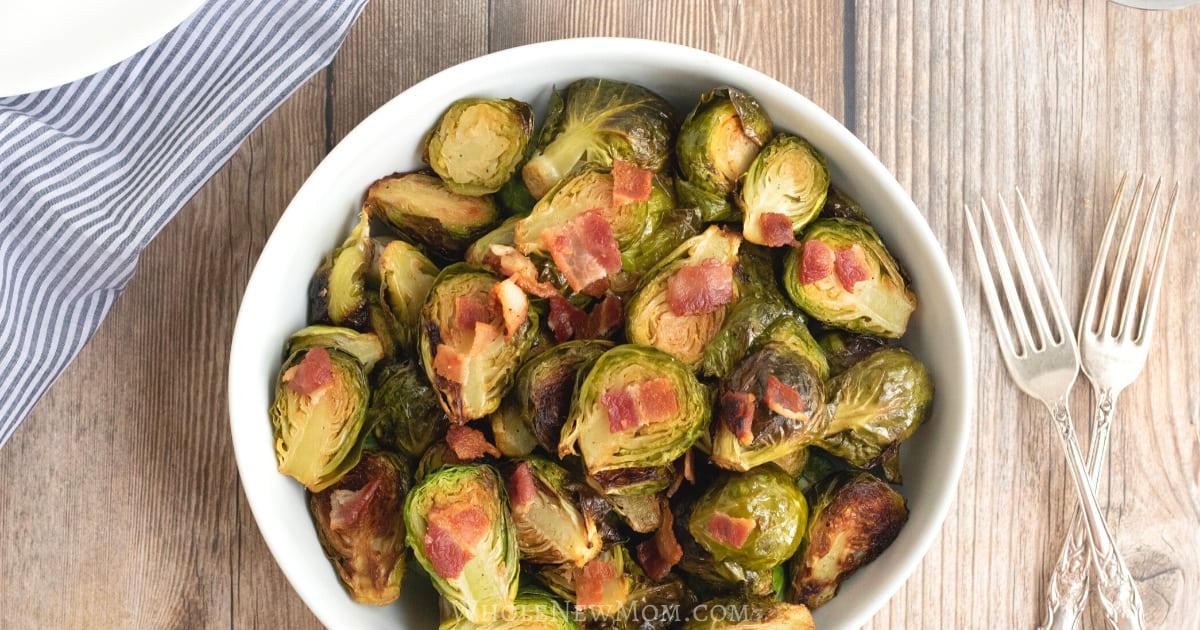 A white bowl filled with roasted Brussels sprouts and pieces of crispy bacon sits on a wooden table. Two silver forks lie beside the bowl, and a blue and white striped cloth is partially visible on the left side.