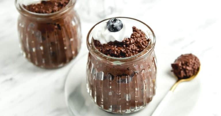 Two glass jars filled with chocolate chia pudding, topped with a dollop of whipped cream and a blueberry, are placed on a white plate. A gold-handled spoon with a small amount of pudding rests beside the plate on a white surface.