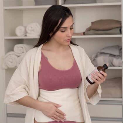 woman wearing Arandi castor oil pack on abdomen while looking at bottle of castor oil.
