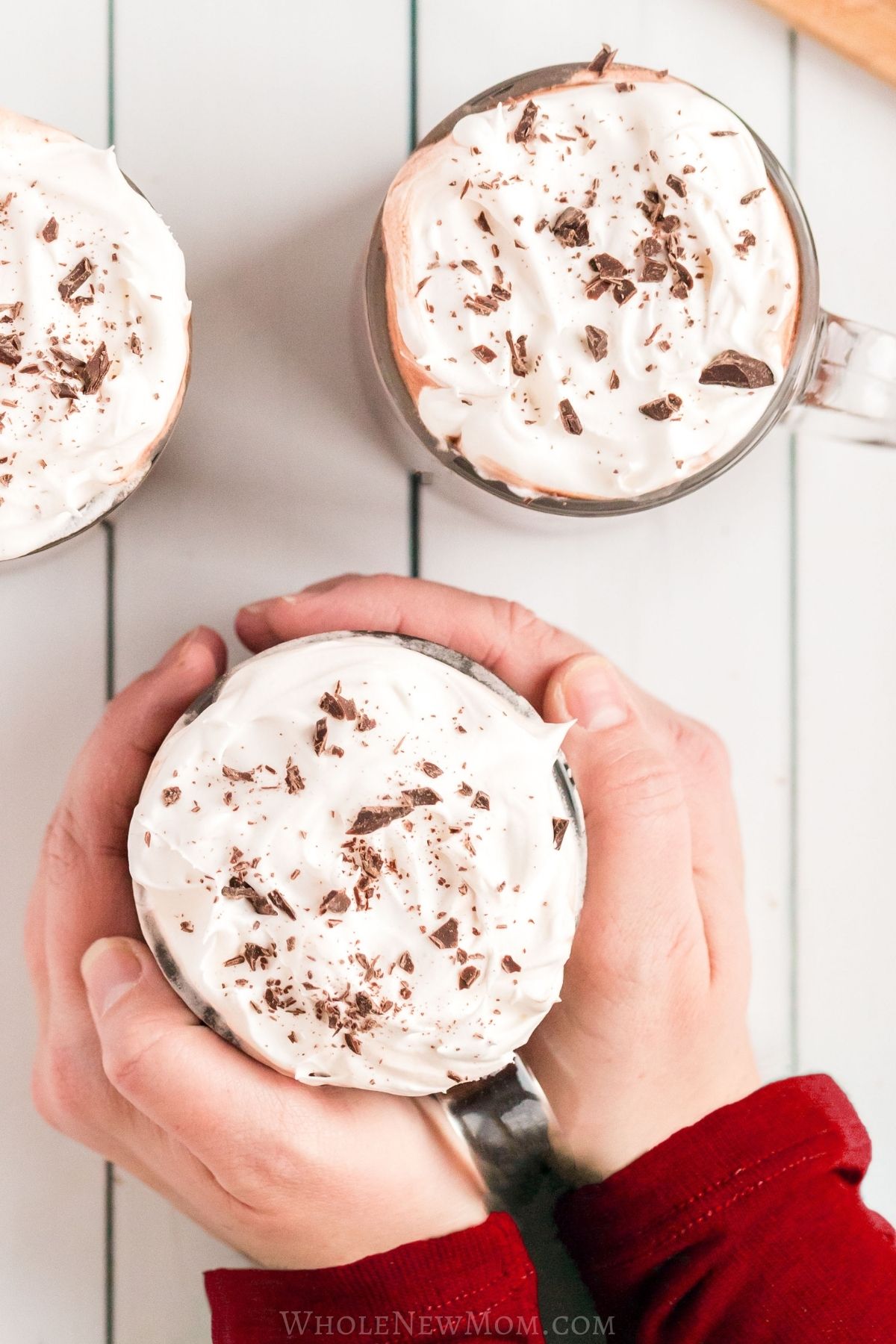 protein hot chocolate with whipped cream and chocolate shavings with hands holding one of the mugs.