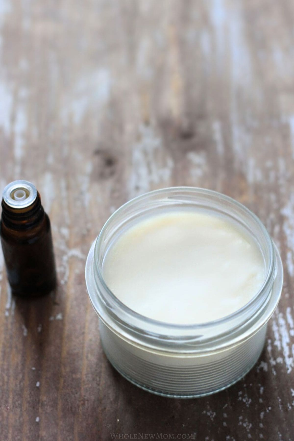 homemade hand lotion in small glass jar on wood table with bottle of essential oils.