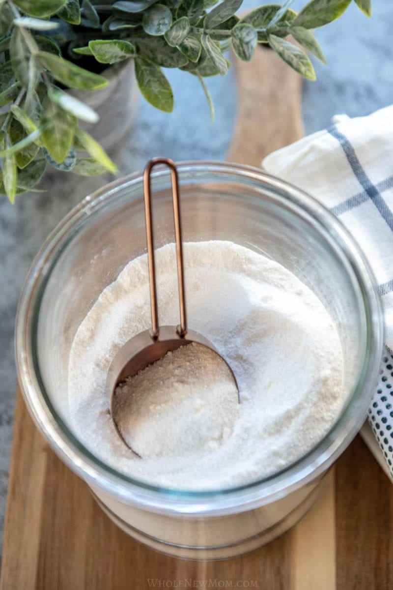 glass container of homemade powdered laundry detergent with metal measuring cup.